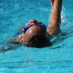 A swimmer performs backstroke in a clear blue pool on a sunny day, wearing blue goggles.