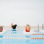 Group exercising on floating mats in an indoor swimming pool setting with a trainer.