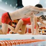Adult men and women engaging in a balance exercise class on poolside boards wearing red swimsuits.