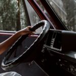 A close-up of a hand gripping a steering wheel in a vintage vehicle, surrounded by forest.