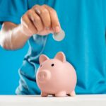 A close-up of an adult's hand dropping a coin into a piggy bank, symbolizing savings and investment.